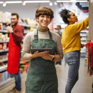 A cheerful woman in a green apron uses a digital tablet, assisting customers in a well-stocked grocery store, reflecting efficient customer service in retail.